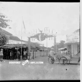 Sebuah gapura bertemakan peringatan Harkitnas dengan seorang abang becak dan beberapa orang di da...