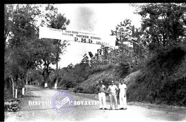 Tiga orang berfoto di bawah spanduk bertuliskan selamat masuk daerah kerja PMD Wlingi, 19 – 8 – 1957