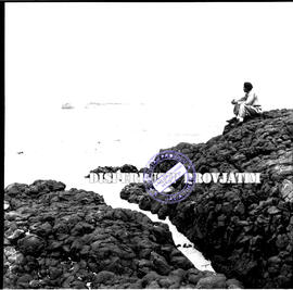 Seorang lelaki tengah duduk di bebatuan (batu konglomerat) sambil menikmati suasana pantai blamba...